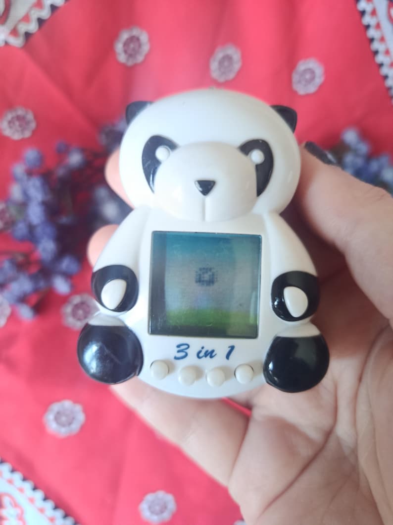 May include: A white and black panda-shaped digital handheld game console featuring a square screen displaying a pixelated graphic. The device has three small white circular buttons below the screen and the text 3 in 1 printed in blue.