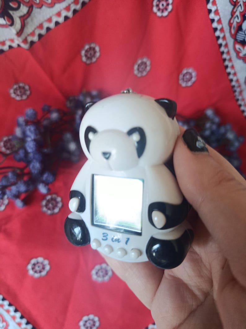 May include: A white and black panda-shaped digital handheld game device with a small square screen. The front of the device features the text 3 in 1 printed in blue above three small white circular buttons. The panda has black ears, eyes, and paws.