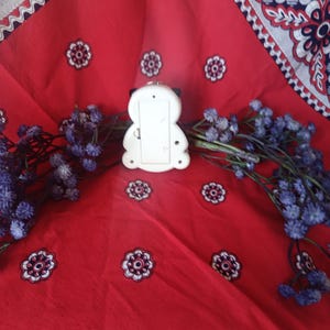 May include: A white plastic object with a rounded, symmetrical shape featuring a rectangular battery compartment cover on the back, positioned centrally between two sprigs of dried purple flowers against a red fabric with a white and blue floral pattern.