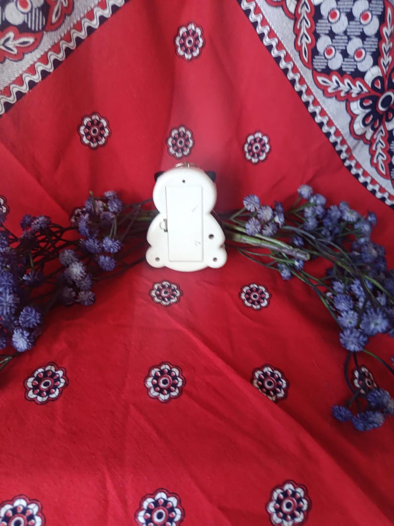 May include: A white plastic object with a rounded, symmetrical shape featuring a rectangular battery compartment cover on the back, positioned centrally between two sprigs of dried purple flowers against a red fabric with a white and blue floral pattern.