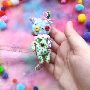 Puede incluir: Una muñeca de tela hecha a mano con una cabeza azul con ojos de botón amarillos y rojos y una boca bordada con una 'X'. La muñeca tiene un cuerpo estampado verde y blanco, un lazo rosa y un cuello con volantes. Pompones coloridos rodean la muñeca.