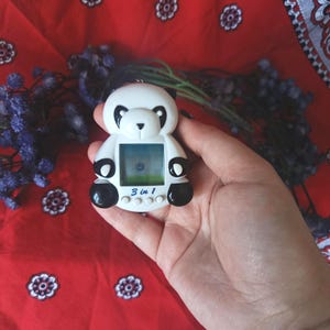 May include: A white and black panda-shaped digital pet device featuring a square screen, three circular control buttons, and the text 3 in 1 printed in blue on the front. The device has black ears, black eye patches, and black paws.