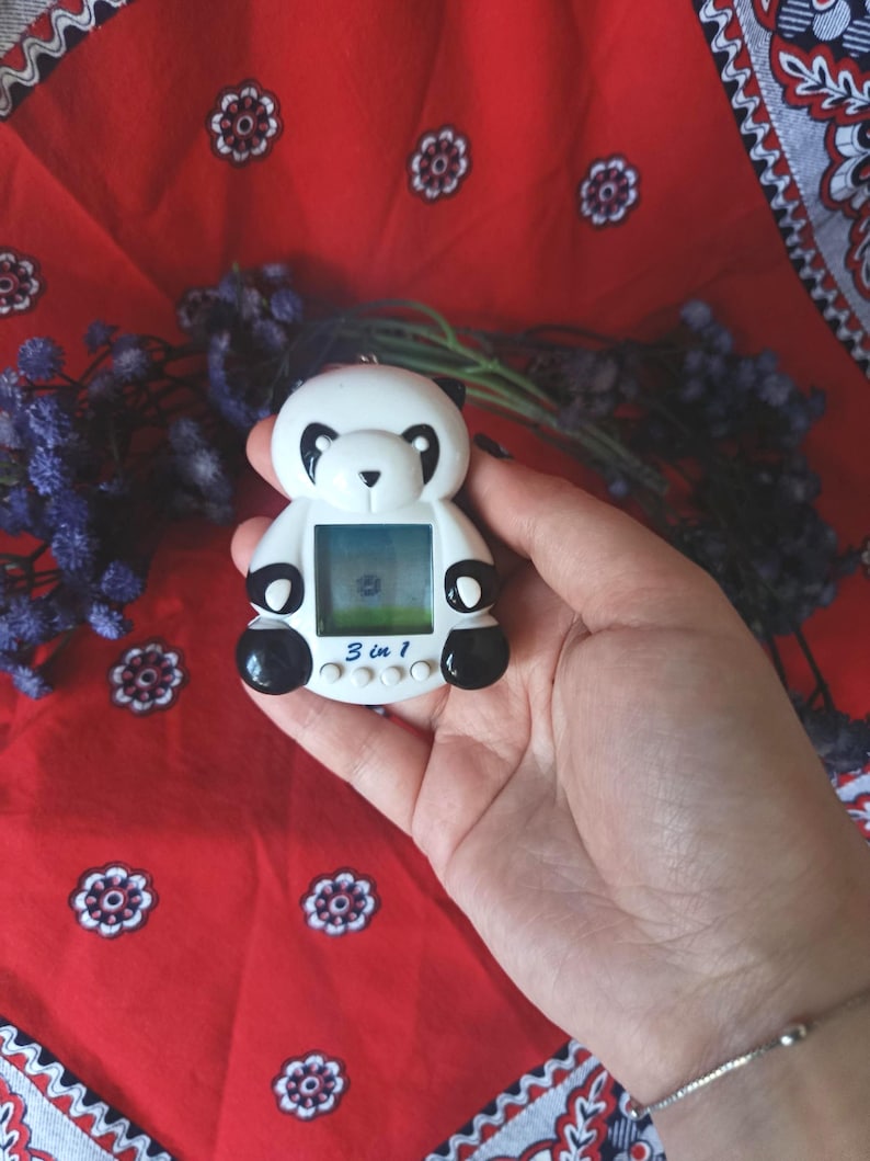 May include: A white and black panda-shaped digital pet device featuring a square screen, three circular control buttons, and the text 3 in 1 printed in blue on the front. The device has black ears, black eye patches, and black paws.