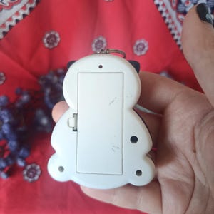 May include: A white plastic electronic handheld game device with a rectangular battery compartment cover on the back. The device features a rounded, asymmetrical shape with small circular screw holes and a metal ring attached at the top edge.