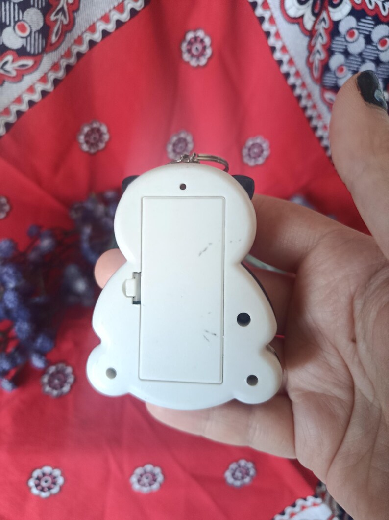 May include: A white plastic electronic handheld game device with a rectangular battery compartment cover on the back. The device features a rounded, asymmetrical shape with small circular screw holes and a metal ring attached at the top edge.