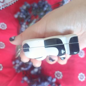 May include: A small, smooth, rectangular object with rounded edges, featuring a high-gloss finish in a black and white cow-print pattern. The item has a thin horizontal seam running across its centre and a small metal loop attached to one side.