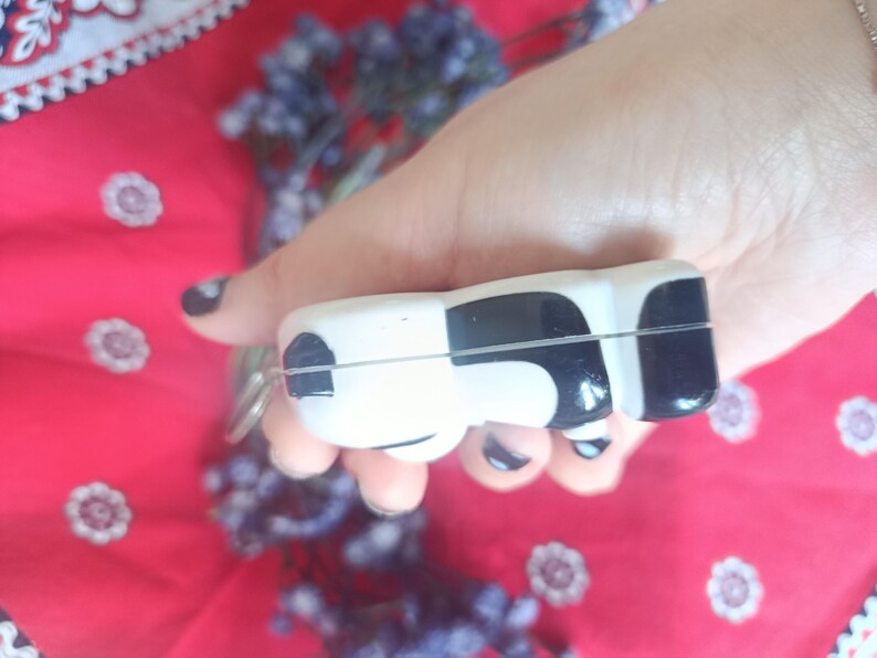 May include: A small, smooth, rectangular object with rounded edges, featuring a high-gloss finish in a black and white cow-print pattern. The item has a thin horizontal seam running across its centre and a small metal loop attached to one side.