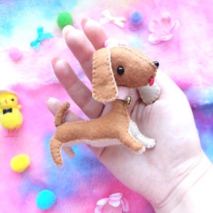 May include: A small, handcrafted felt dachshund plush toy in shades of brown and cream. The dog features black bead eyes, a tiny red tongue, and a small gold-colored bell attached to a light pink collar around its neck with visible stitching.
