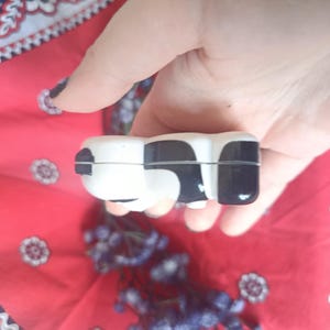 May include: A small, smooth, ceramic trinket box shaped like a cow, featuring a glossy white base with distinct black patches. The object has a horizontal seam running through the middle, indicating a lid that opens to reveal a hollow interior space.