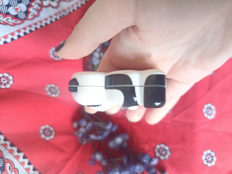 May include: A small, smooth, ceramic trinket box shaped like a cow, featuring a glossy white base with distinct black patches. The object has a horizontal seam running through the middle, indicating a lid that opens to reveal a hollow interior space.