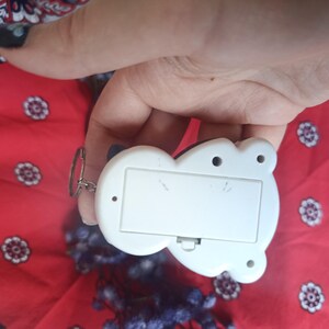 May include: A white plastic electronic keyring device with a rectangular battery compartment cover on the back, featuring a small metal ring attached to the side for keys, presented against a red fabric background with a white floral pattern.