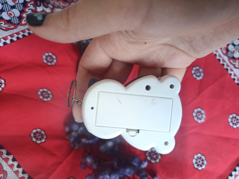 May include: A white plastic electronic keyring device with a rectangular battery compartment cover on the back, featuring a small metal ring attached to the side for keys, presented against a red fabric background with a white floral pattern.