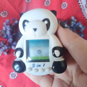 May include: A white and black panda-shaped electronic handheld game console with a digital screen. The device features a small display showing a landscape, four circular control buttons at the bottom, and the text 3 in 1 printed on the front.