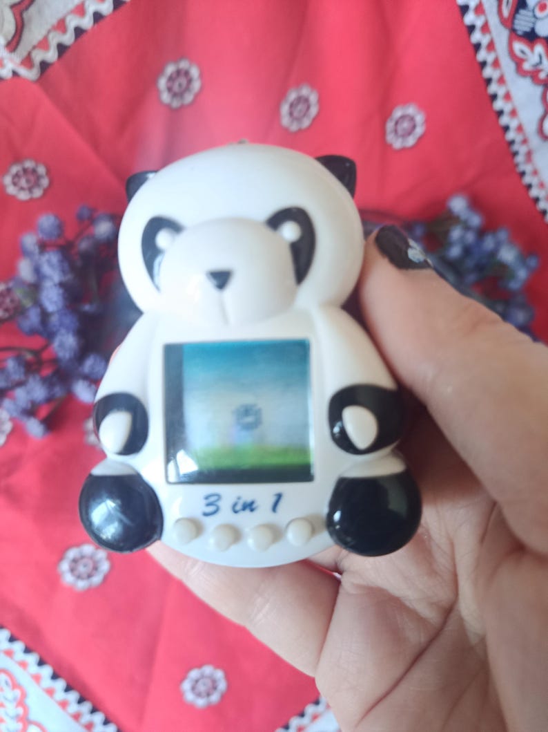 May include: A white and black panda-shaped electronic handheld game console with a digital screen. The device features a small display showing a landscape, four circular control buttons at the bottom, and the text 3 in 1 printed on the front.