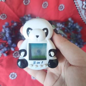May include: A white and black panda-shaped digital handheld game console featuring a small square screen, four circular control buttons below the display, and the text 3 in 1 printed in blue on the front surface of the plastic device.