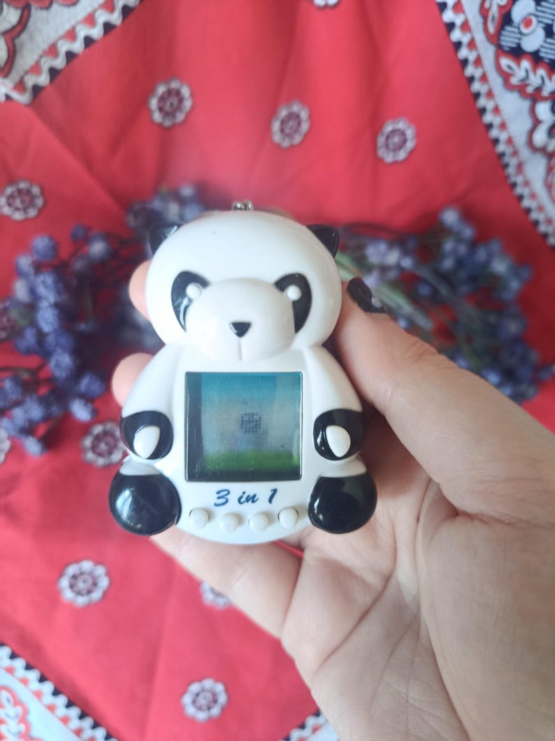 May include: A white and black panda-shaped digital handheld game console featuring a small square screen, four circular control buttons below the display, and the text 3 in 1 printed in blue on the front surface of the plastic device.