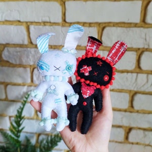 Puede incluir: Dos muñecos de peluche de conejito hechos a mano. Uno es blanco con detalles azules, el otro es negro con detalles rojos. El conejito blanco tiene orejas a rayas azules y un lazo azul. El conejito negro tiene orejas a cuadros rojas y blancas y un corazón rojo.