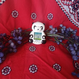 May include: A white and black panda-shaped digital pet toy with a small screen displaying a graphic. The device features the text 3 in 1 printed below the screen and includes three circular buttons for navigation and control of the electronic game.
