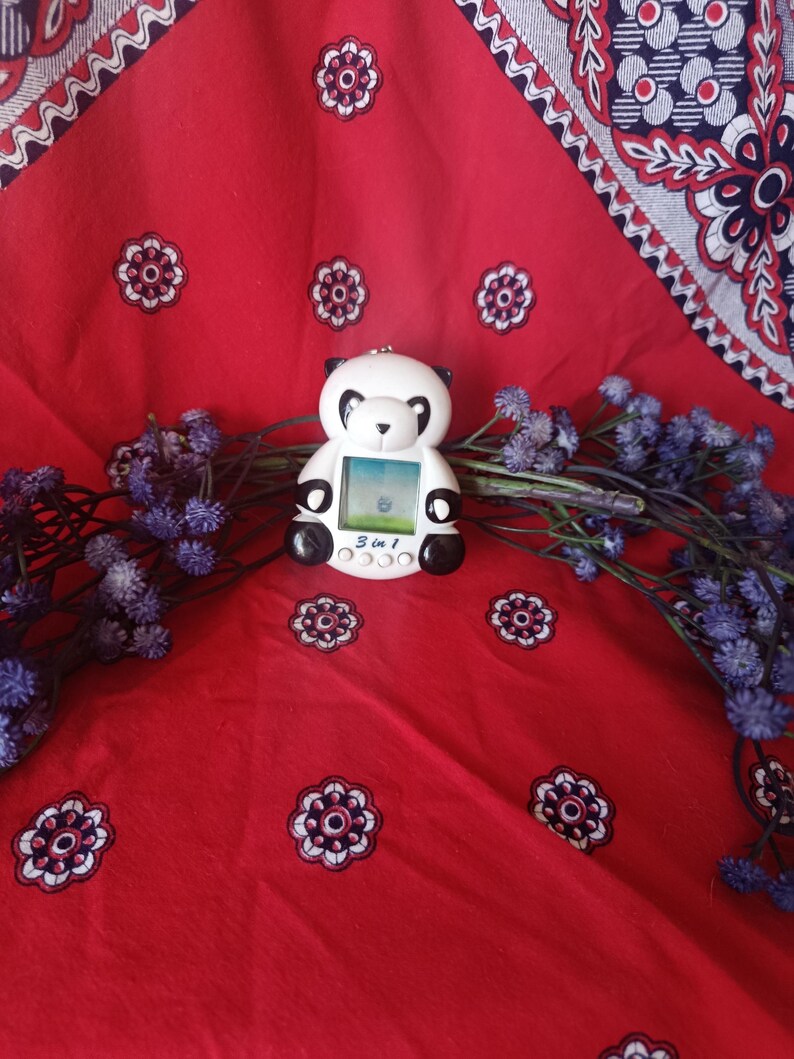 May include: A white and black panda-shaped digital pet toy with a small screen displaying a graphic. The device features the text 3 in 1 printed below the screen and includes three circular buttons for navigation and control of the electronic game.