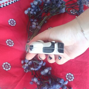 May include: A small, rectangular, white and black cow-patterned protective case for wireless earbuds. The case features a smooth, glossy finish with a distinct black and white spotted design covering the exterior surface of the compact device.