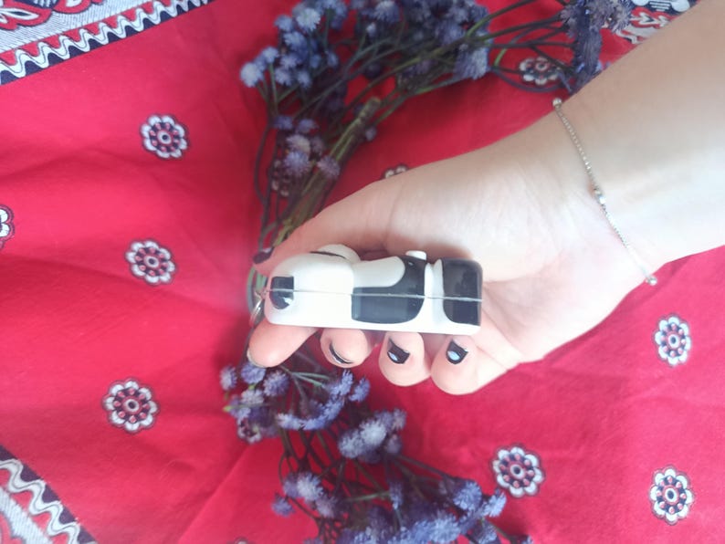 May include: A small, rectangular, white and black cow-patterned protective case for wireless earbuds. The case features a smooth, glossy finish with a distinct black and white spotted design covering the exterior surface of the compact device.