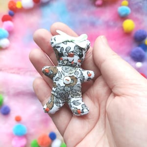 Puede incluir: Pequeña muñeca de oso de peluche hecha a mano con un estampado floral en tonos grises y marrones. La muñeca tiene un lazo verde claro y un botón. La muñeca se sostiene en una mano, con pompones de colores en el fondo.