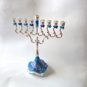 May include: Silver menorah with nine candle holders, each with a blue and turquoise decorative band. The base of the menorah is decorated with a blue and pink floral pattern.