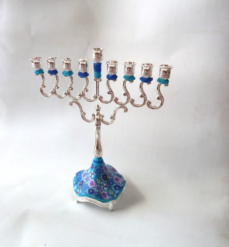 May include: Silver menorah with nine candle holders, each with a blue and turquoise decorative band. The base of the menorah is decorated with a blue and pink floral pattern.
