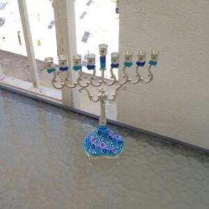 May include: A silver menorah with blue and white floral design on the base. The menorah has nine candle holders and is decorated with blue and white ribbons.