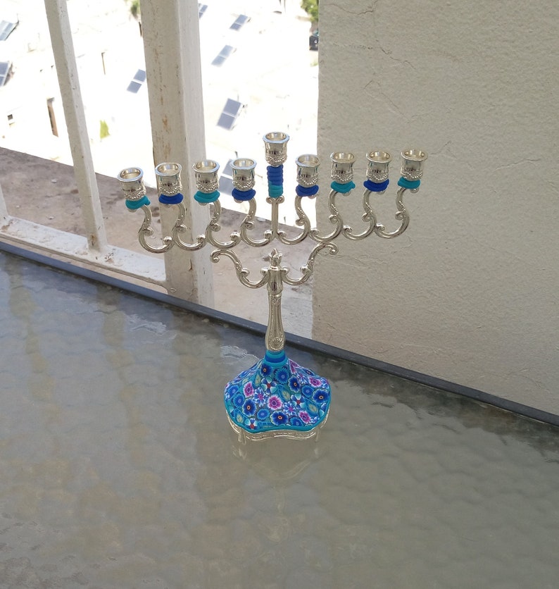 May include: A silver menorah with blue and white floral design on the base. The menorah has nine candle holders and is decorated with blue and white ribbons.