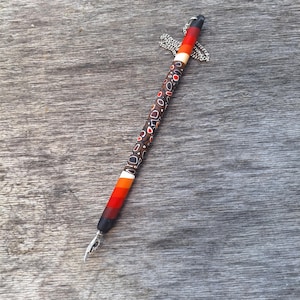 Può includere: Una penna decorativa con pennino argentato e fusto fantasia. Il fusto presenta un disegno a macchie e strisce marroni, arancioni e bianche. La penna ha una sfumatura di colori dal nero al rosso all'arancione. Una catena d'argento è attaccata in alto.