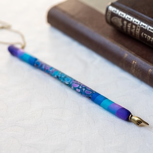 Może przedstawiać: Kolorowe pióro wieczne ze złotą stalówką, z żywym wzorem w odcieniach niebieskiego, fioletowego i turkusowego. Pióro leży na białej powierzchni, z brązową książką w tle. Wzór pióra zawiera wzór wirów i kropek.