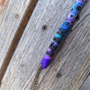 May include: A colorful, handmade pendant with the name "LEAHA" in black letters. The pendant features a pattern of blue, purple, and black swirls and stripes. A small gold chain is attached to the bottom of the pendant.