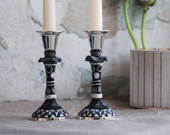 May include: Two black and white ceramic candle holders with silver bases and white candles. The candle holders have a geometric pattern and are on a light brown surface.