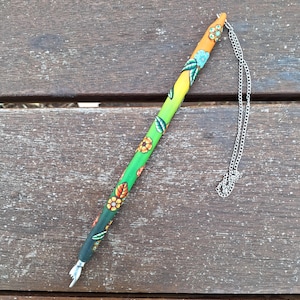 May include: A decorative pen with a floral design. The pen has a colour gradient from orange to green, and is decorated with flowers and leaves. A silver chain is attached to the pen.