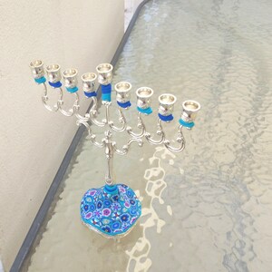 May include: Silver menorah with blue and turquoise decorative accents. The base of the menorah is decorated with a floral pattern in shades of blue, pink, and white.