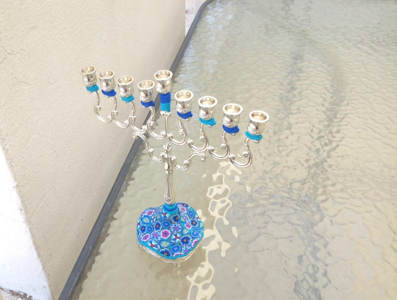 May include: Silver menorah with blue and turquoise decorative accents. The base of the menorah is decorated with a floral pattern in shades of blue, pink, and white.