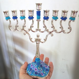 May include: Silver menorah with blue and turquoise decorative accents. The base is decorated with a floral pattern in shades of blue, turquoise, and purple.