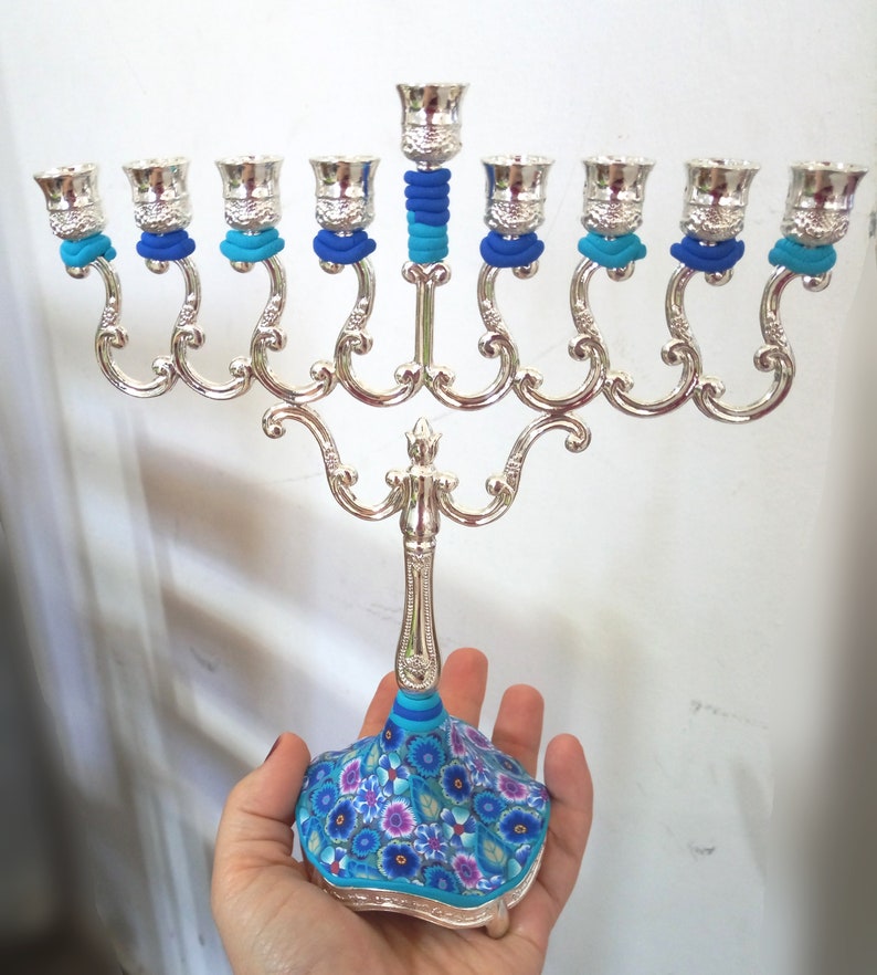 May include: Silver menorah with blue and turquoise decorative accents. The base is decorated with a floral pattern in shades of blue, turquoise, and purple.