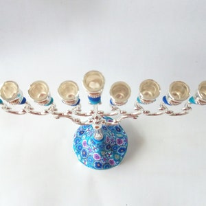 May include: Silver menorah with a blue and white floral base. The menorah has nine candle holders and is decorated with blue and silver accents.