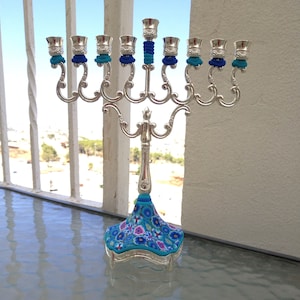 May include: Silver menorah with blue and white floral design on the base. The menorah has nine candle holders and is decorated with blue and white trim.