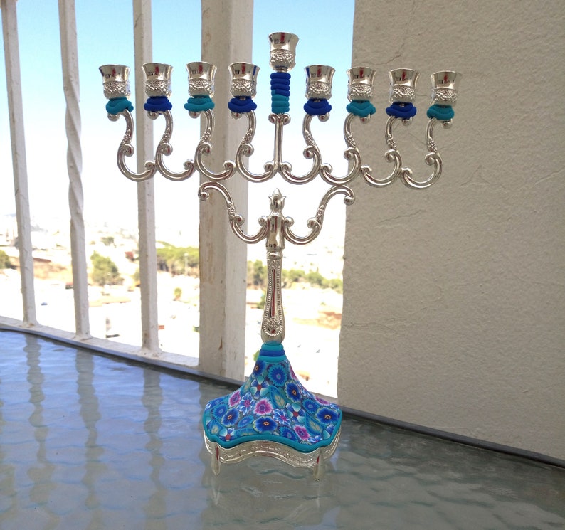 May include: Silver menorah with blue and white floral design on the base. The menorah has nine candle holders and is decorated with blue and white trim.