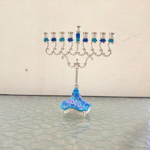 May include: A silver menorah with nine candle holders, each with a blue and turquoise glass candle cup. The menorah has a floral patterned base with blue, pink, and white flowers.