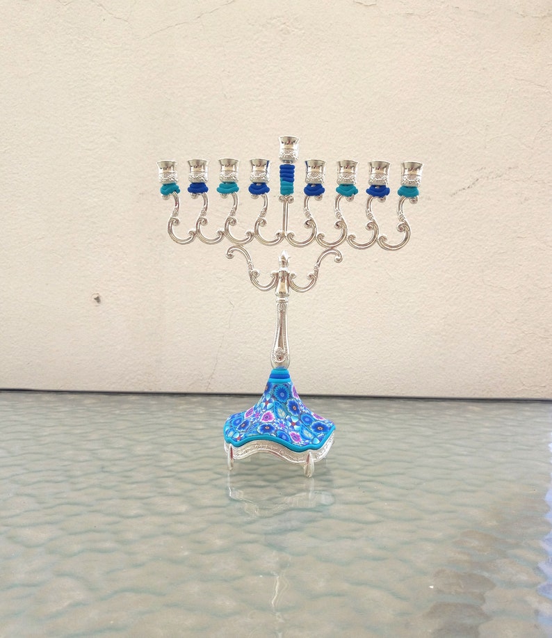 May include: A silver menorah with nine candle holders, each with a blue and turquoise glass candle cup. The menorah has a floral patterned base with blue, pink, and white flowers.