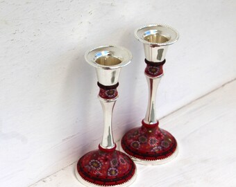 May include: A pair of silver candle holders with red and black floral designs. The candle holders are tall and slender, with a round base.