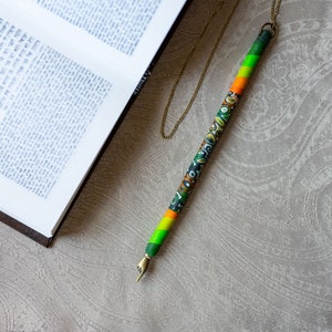 May include: A colorful pen-shaped pendant with a gold-colored chain. The pen features a green, orange, and patterned design. The pen's tip is shaped like a pointing finger. An open book with Hebrew text is in the background.