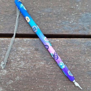 Może przedstawiać: Dekoracyjne pióro maczane z gradientem od niebieskiego do fioletowego, ozdobione wzorami kwiatowymi i liściastymi. Pióro posiada srebrną stalówkę i pasujący łańcuszek. Pióro ma około 18 cm długości.