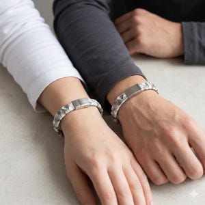 以下が含まれることがあります： テクスチャード加工のパターンと長方形の留め金が付いた、お揃いのシルバーと白のブレスレットが2つ。 ブレスレットは手首に着用されており、一人は白い長袖シャツを、もう一人は灰色の長袖シャツを着ています。