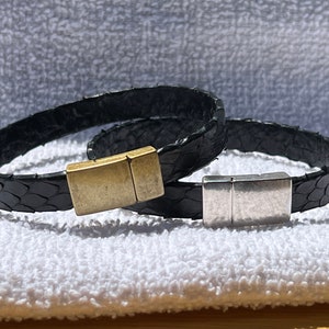 Peut inclure: Deux bracelets en cuir noir avec fermoirs magnétiques. Un fermoir est doré et l'autre est argenté.