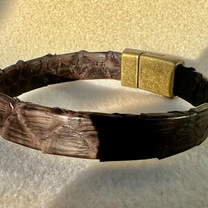 Pu&ograve; includere: Un braccialetto in pelle marrone con un motivo strutturato a forma di serpente. Il braccialetto presenta una chiusura magnetica rettangolare color oro. Il braccialetto &egrave; largo circa 2 cm.